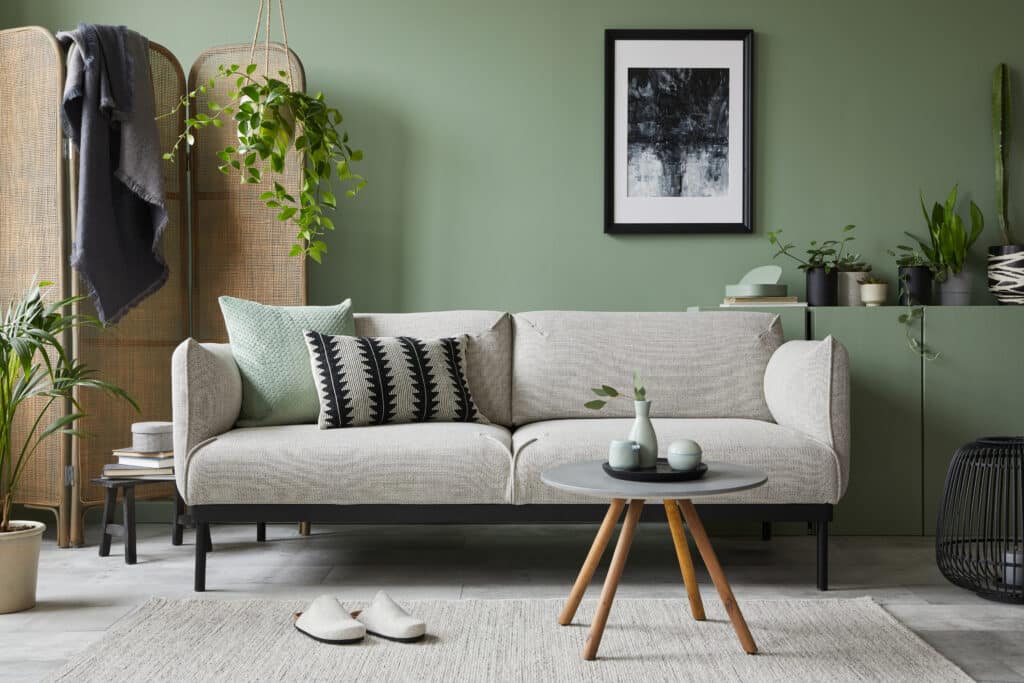 Stylish composition of modern living room interior. Mock up poster frame, modern sofa, folding screen, plants and personal accessories. Sage green wall. Home staging. Template. Copy space.